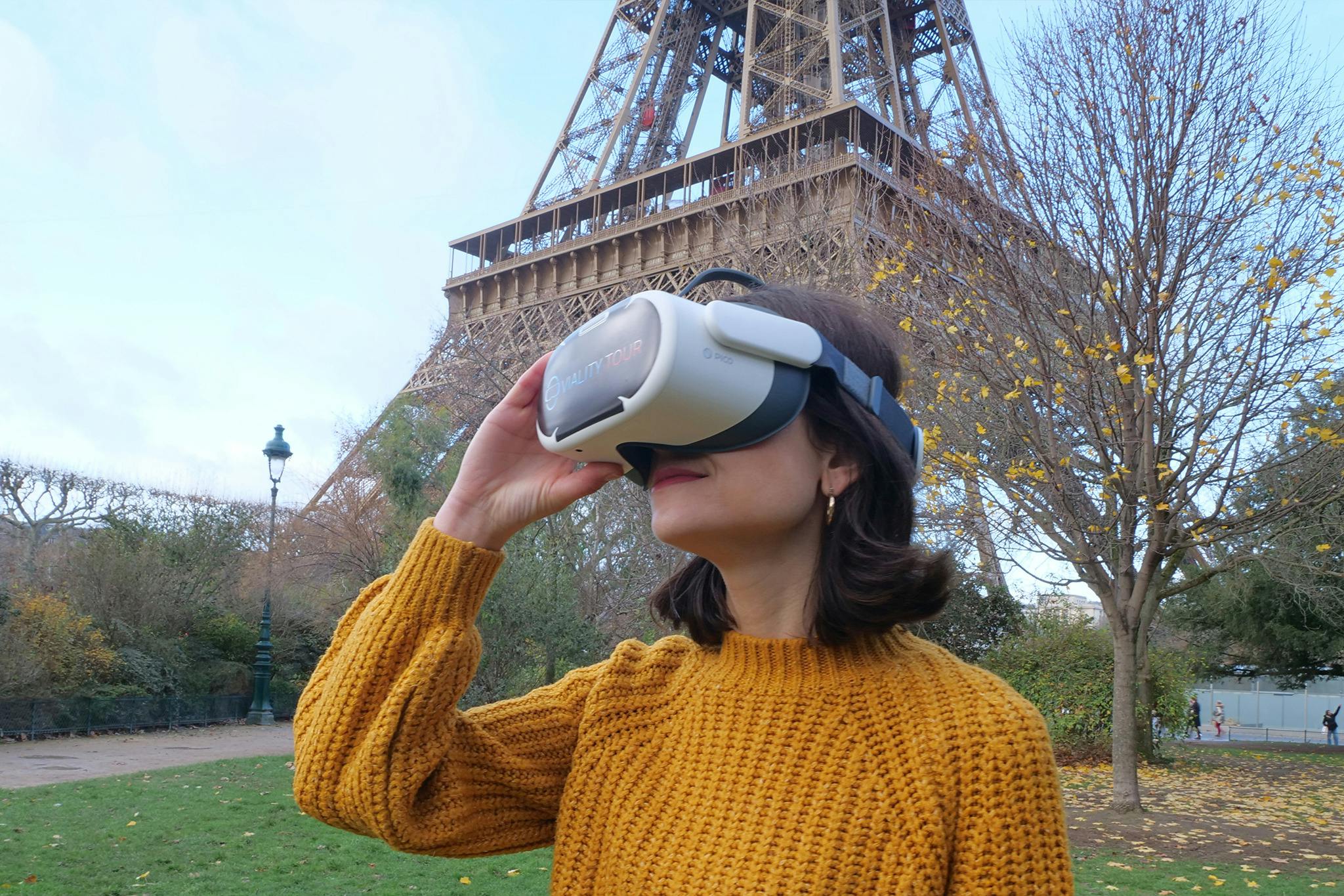Una persona con un maglione giallo utilizza una cuffia per la realtà virtuale vicino alla Torre Eiffel in un parco in una giornata limpida.