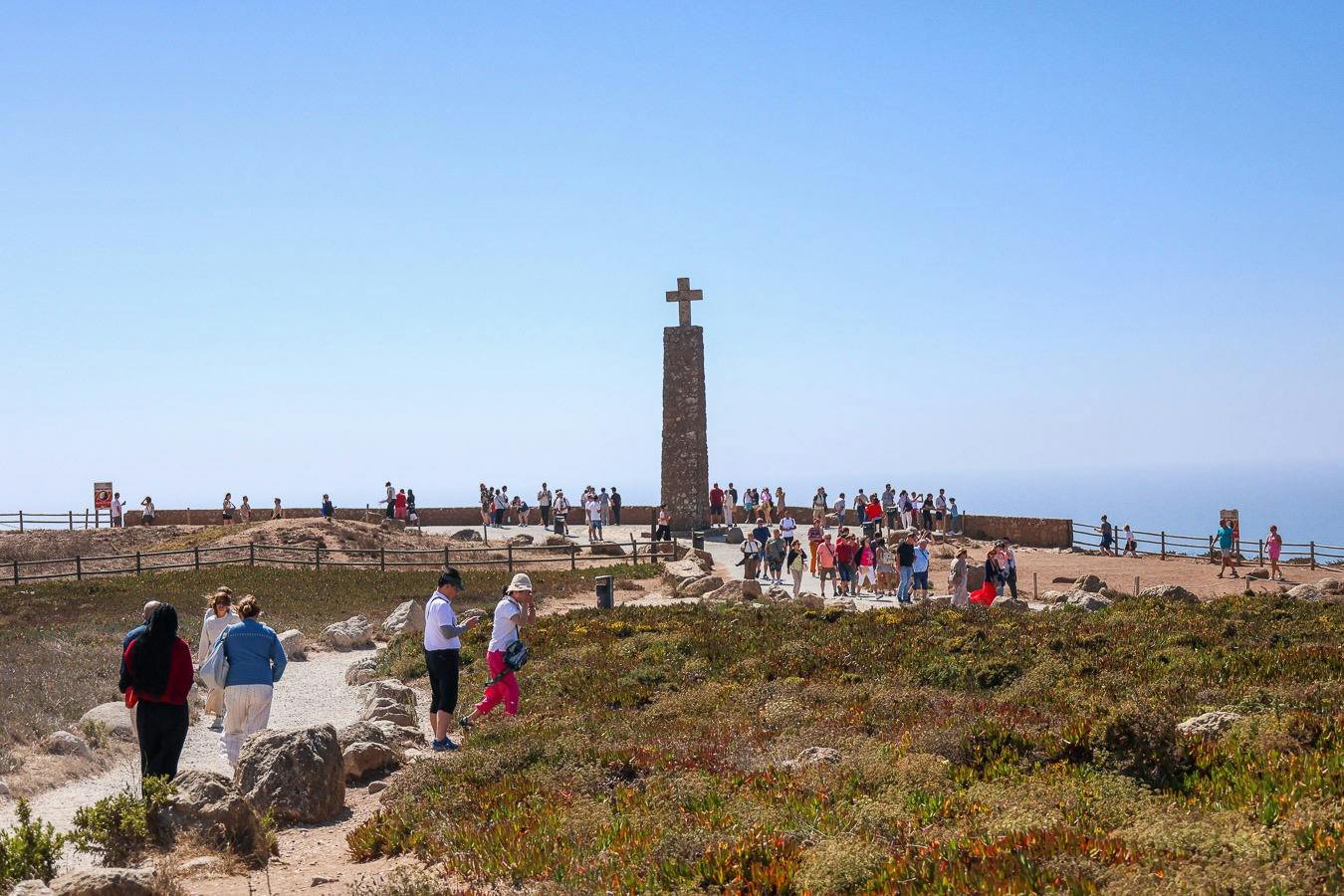 A group of people gathered near a stone monument with a cross on top, set in a coastal area under a clear blue sky.