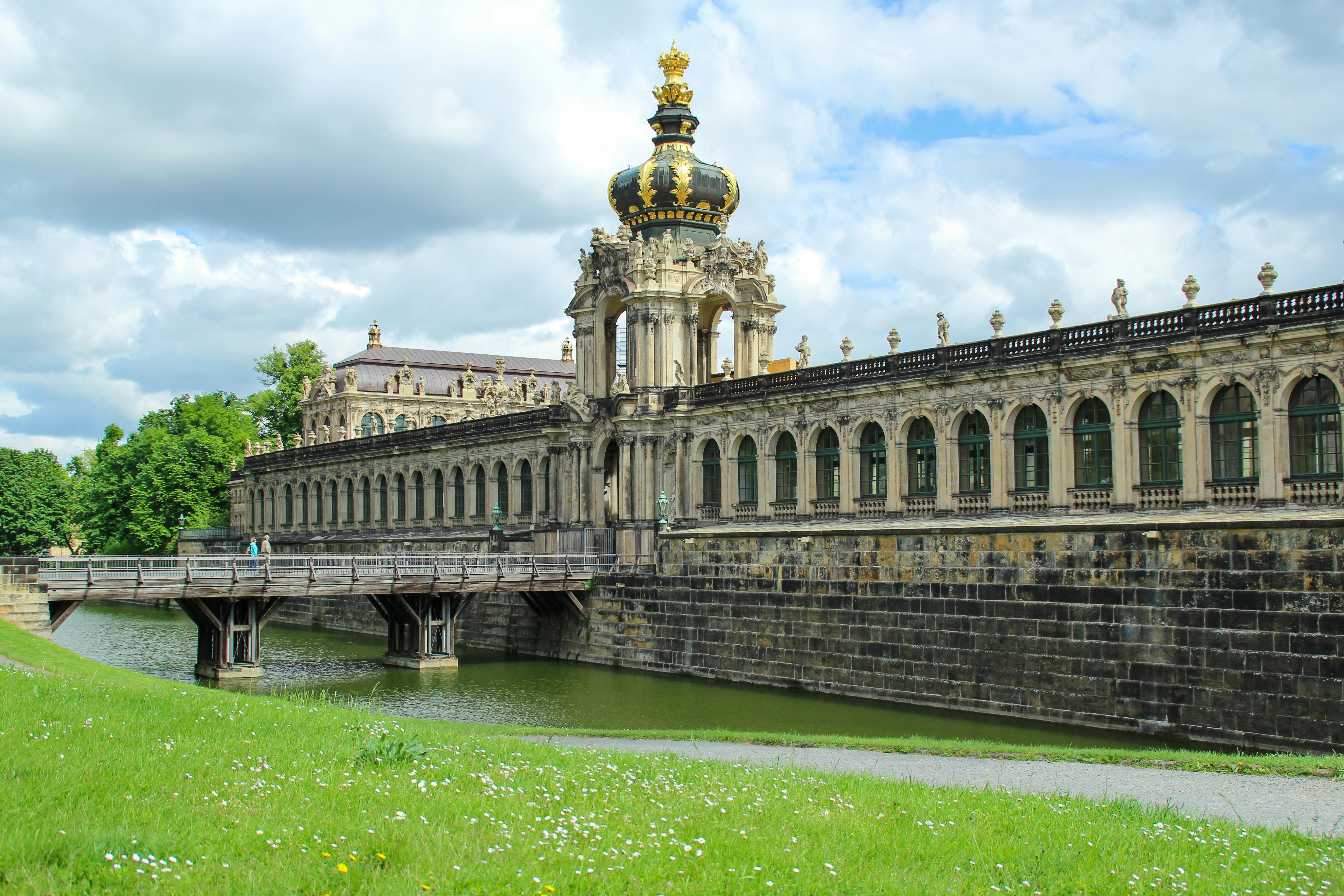Long, ornate Baroque building with an elaborately decorated crown atop, a stone bridge over a moat, and a grassy area in the foreground.