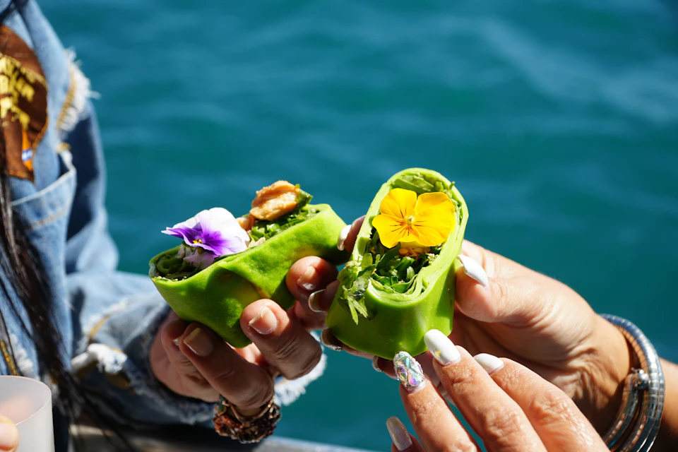 Two hands holding green wraps with edible flowers and greens inside, against a blurred blue background, likely water.