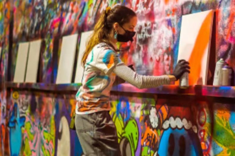 En person, der bærer maske og handsker, sprayer et lærred mod en graffiti-beklædt væg med flere blanke lærreder i nærheden.