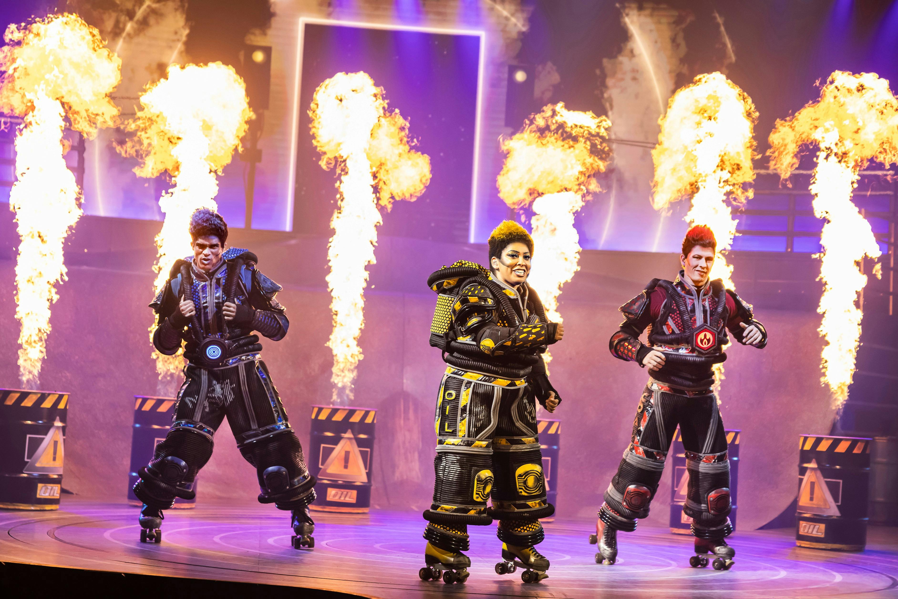 两位穿着未来服装的表演者在舞台上滑轮滑冰，背景中有火焰爆发和霓虹灯效果。