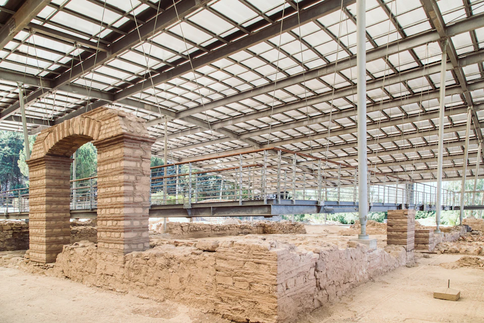 Ancient ruins with brick structures under a modern metal and glass roof, with a raised walkway in the background.