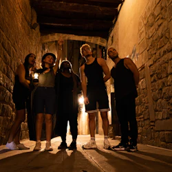 Five people standing in a narrow alley, with one person pointing upwards. The scene is dimly lit, creating a mysterious atmosphere.