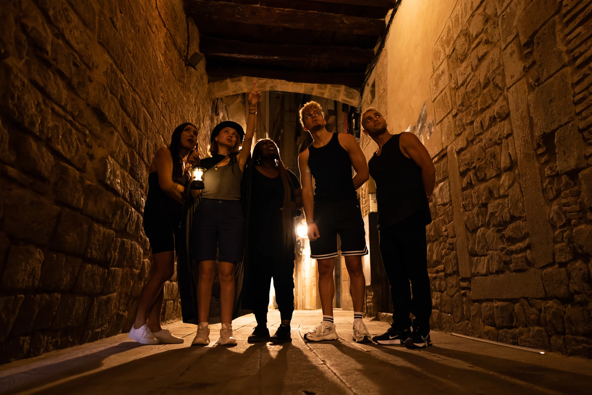 Five people standing in a narrow alley, with one person pointing upwards. The scene is dimly lit, creating a mysterious atmosphere.