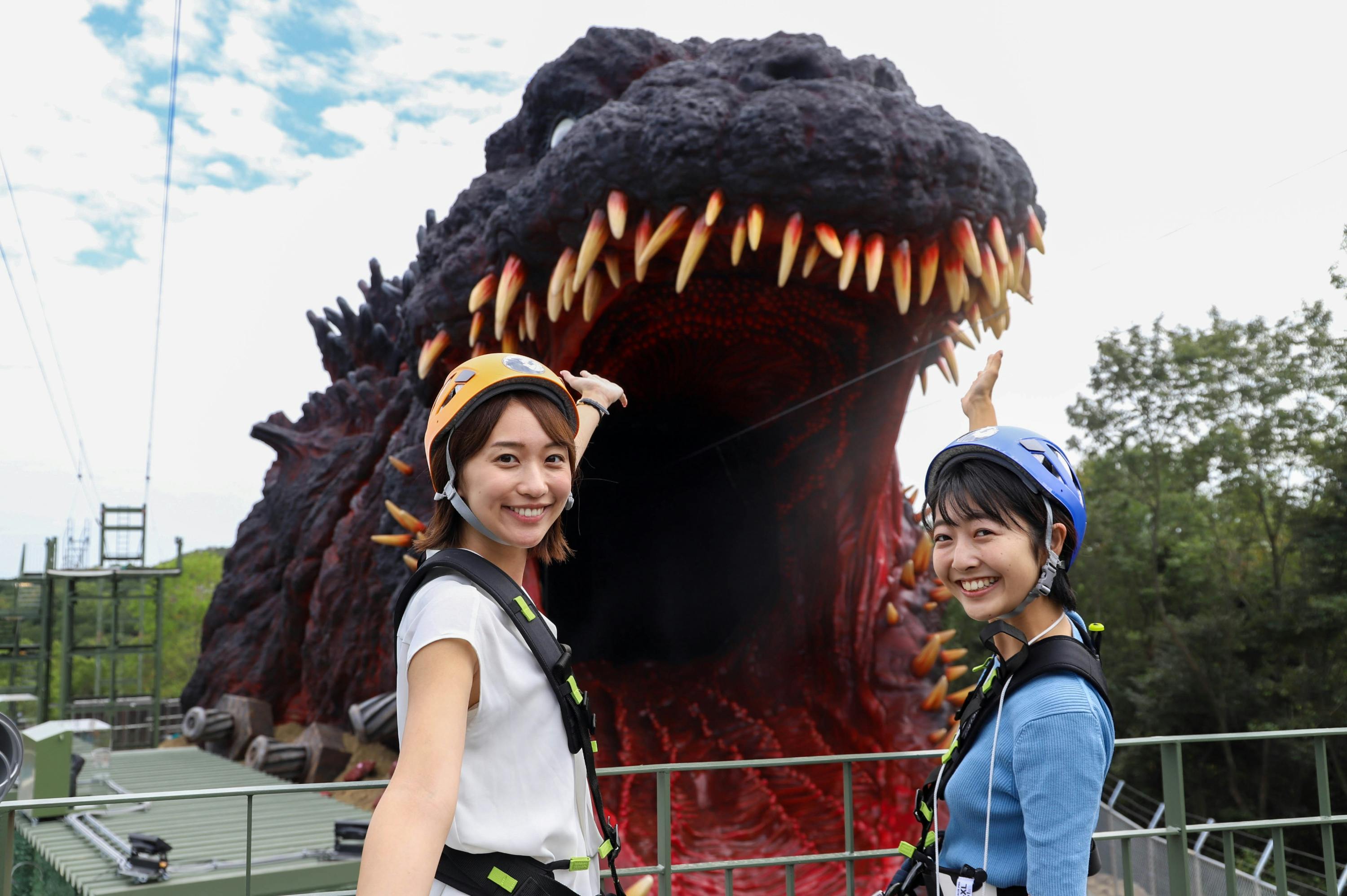 Two women wearing helmets and harnesses pose in front of a large dinosaur-like statue with an open mouth.