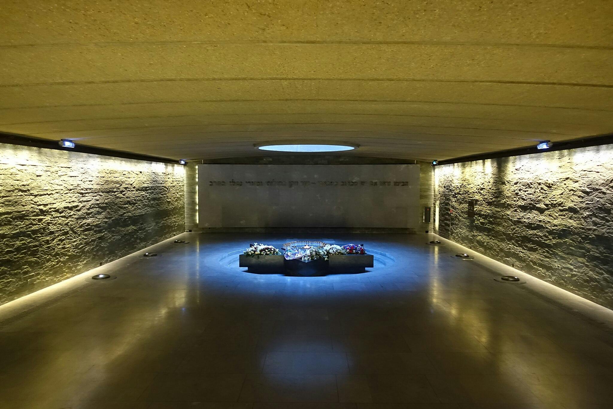 A solemn, dimly-lit room with stone walls houses a circular monument adorned with floral wreaths under a central skylight.