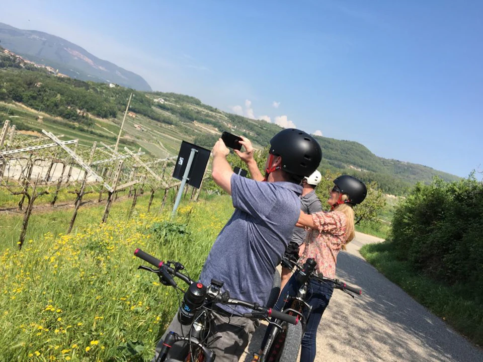 Three people wearing helmets stand with bicycles on a path, one taking a photo, overlooking a hilly, green landscape.