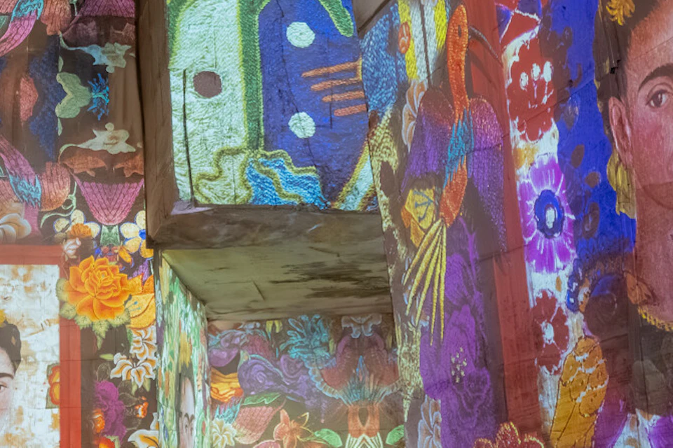 Colorful digital projections of Frida Kahlo's artwork on large walls, with people silhouetted in the foreground.