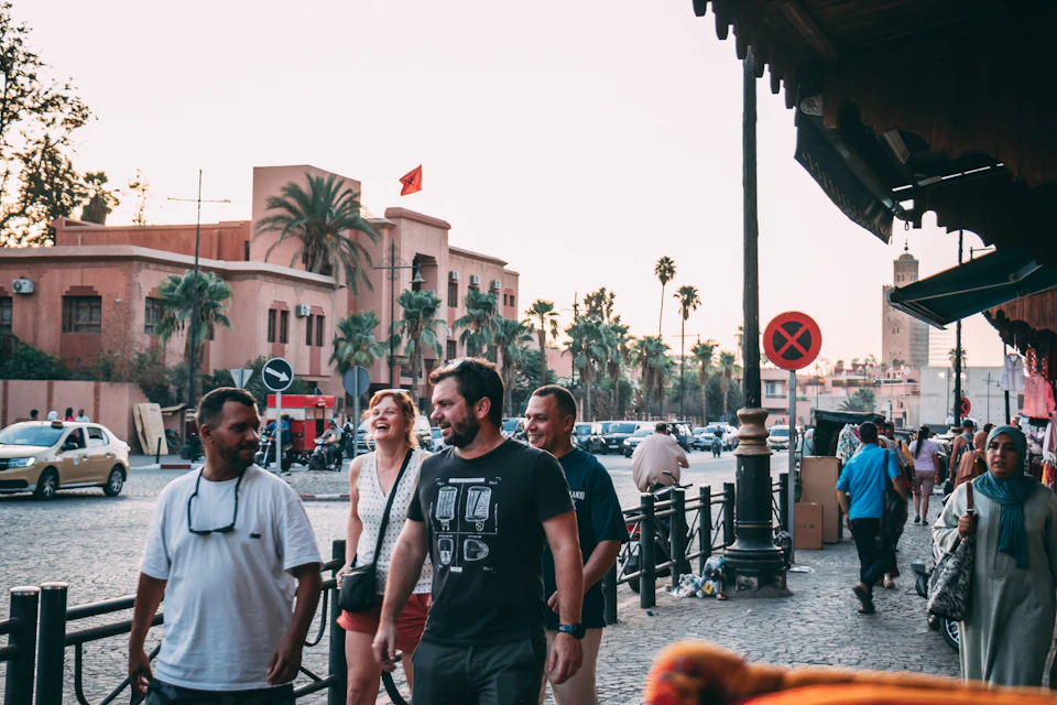 A group of four people walk and laugh on a city street with palm trees and a beige building with a red flag in the background.