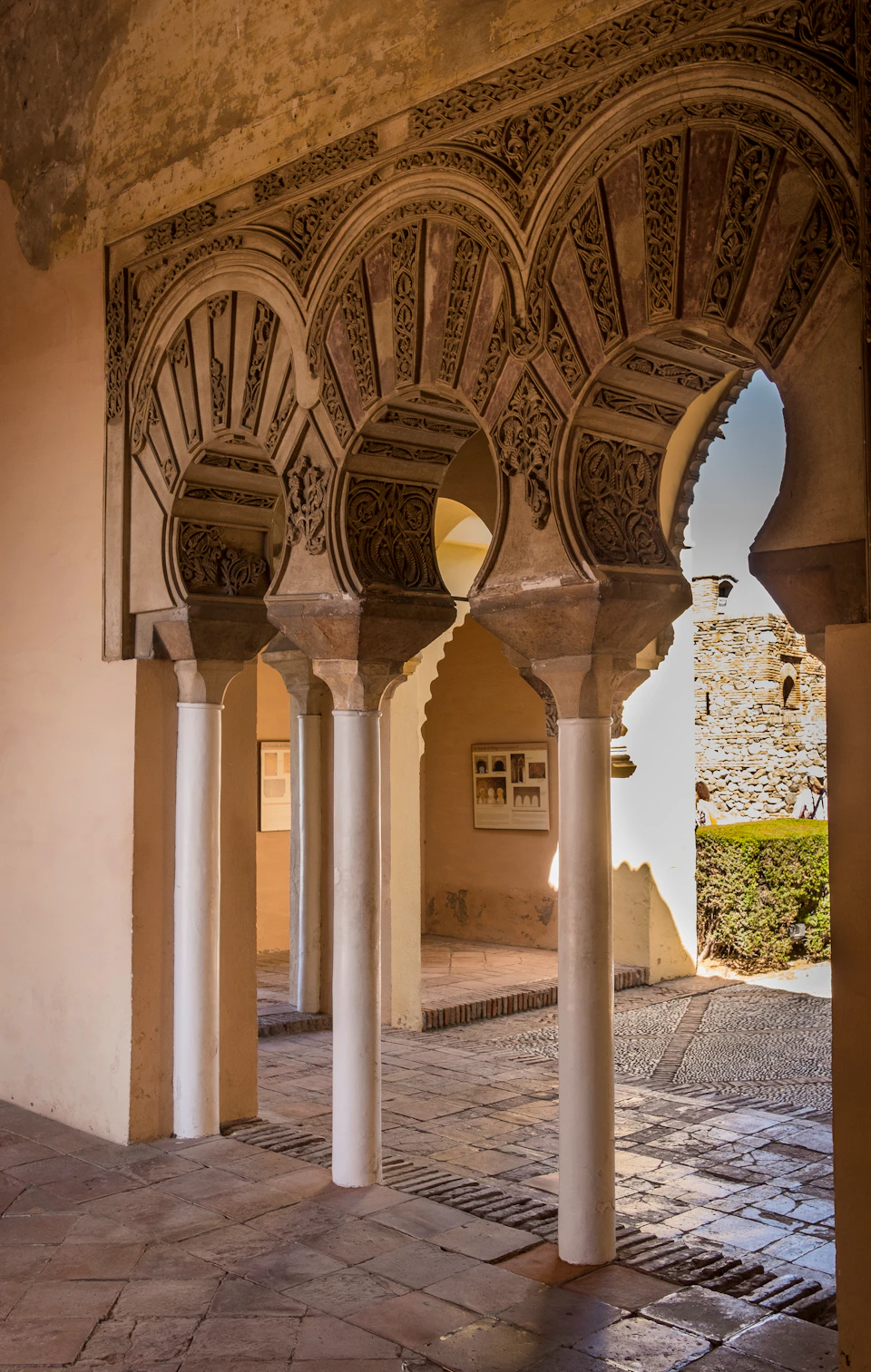 Intricately carved arches supported by columns, with sunlight illuminating a courtyard and a stone wall in the background.