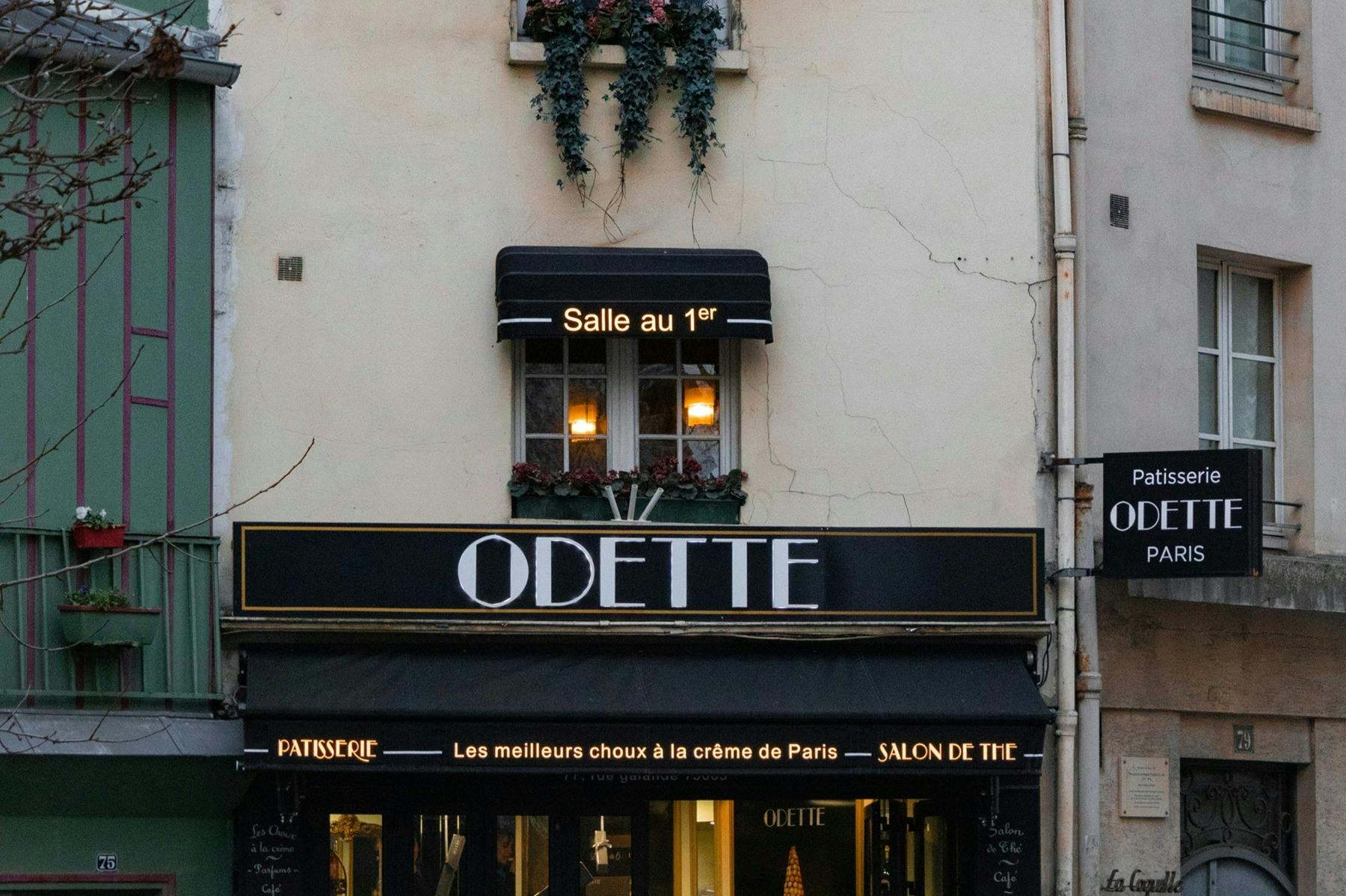 The facade of a patisserie named Odette, featuring decorative signs and lights, with window boxes of flowers above.