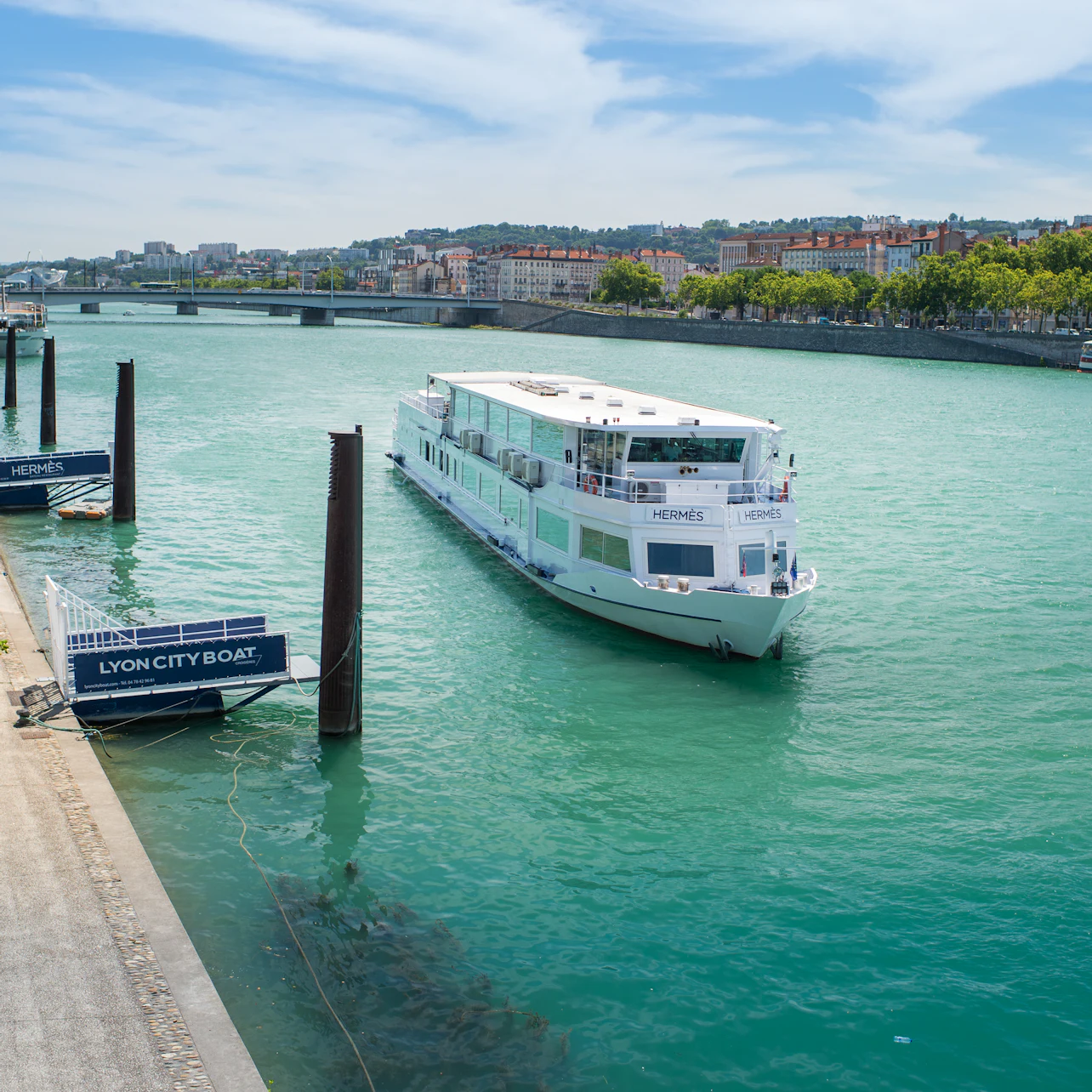 A white tour boat labeled "HERMÈS" approaches a dock on a wide river with a cityscape and bridge in the background.
