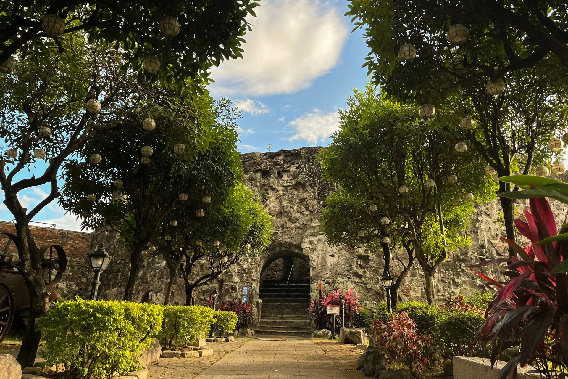 A stone archway surrounded by trees and greenery under a blue sky with scattered clouds.