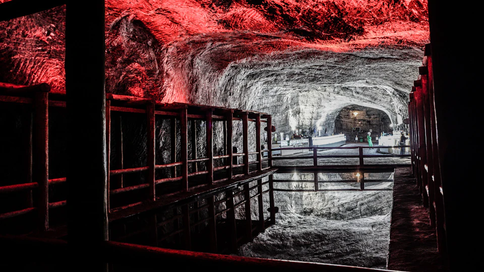 Underground cavern with red lighting on the ceiling, a wooden railing, and a still water reflection. People are visible in the background.