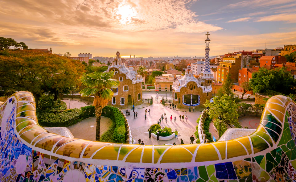 People walking in a colorful park with two uniquely designed buildings, lush greenery, and a vibrant sunset sky in the background.