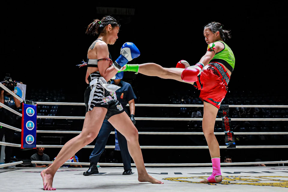 Two female Muay Thai fighters in a ring, one delivering a high kick. Both wear gloves and athletic gear. Referee in the background.