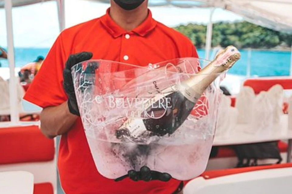 A person wearing a red shirt and black gloves holding a clear ice bucket with a champagne bottle inside.