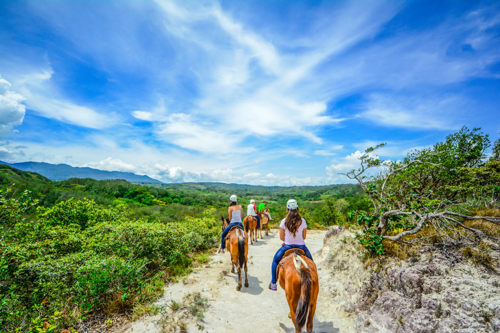 People riding horses on a dirt trail through a green, hilly landscape under a bright blue, partly cloudy sky.