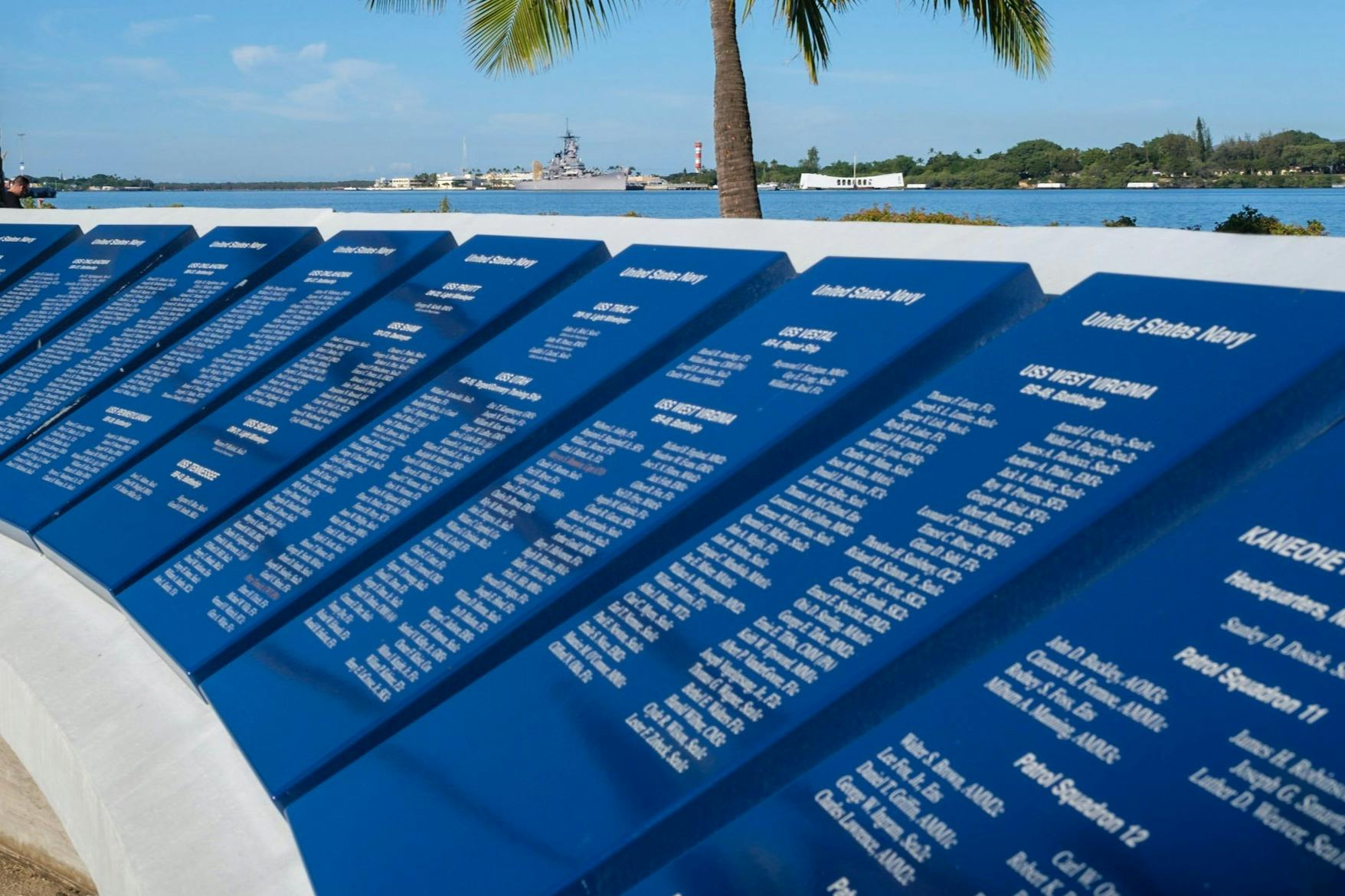 Blue memorial plaques with names, a palm tree, and a battleship in the background near a waterfront.
