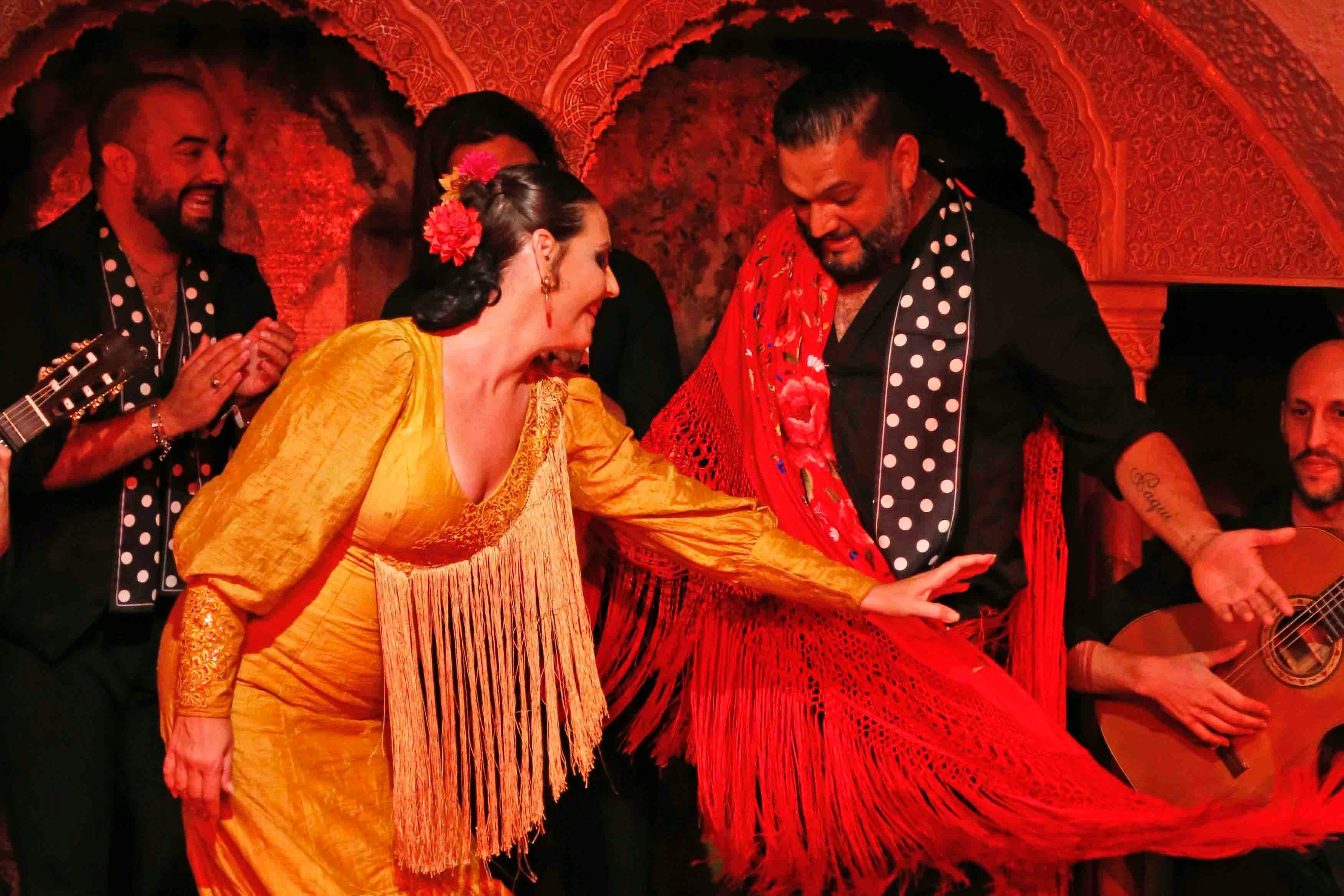 Kobieta w żółtej sukience flamenco i mężczyzna w czarnej koszuli z czerwonymi akcentami wykonują taniec w żywej scenerii.
