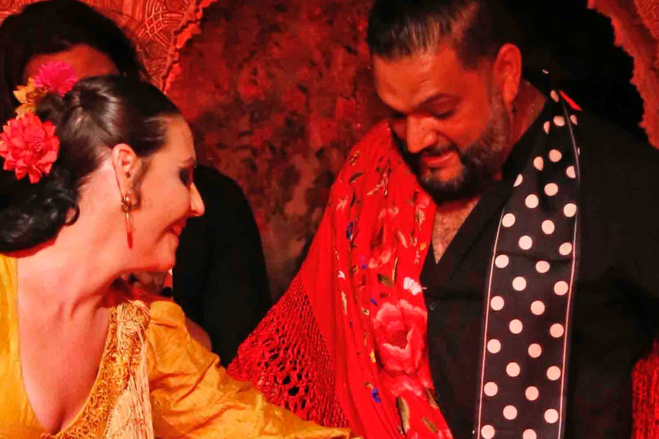 A woman in a yellow flamenco dress and a man in a black shirt with red accents perform a dance in a vibrant setting.