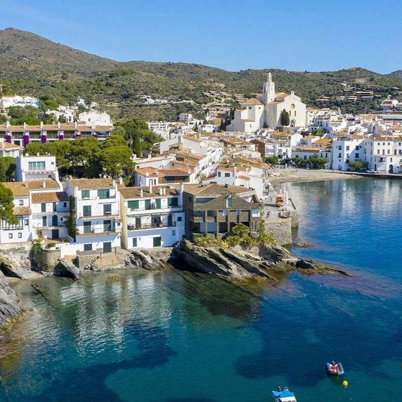 A coastal town with white buildings and a prominent church near a blue bay, surrounded by hills under a clear sky.