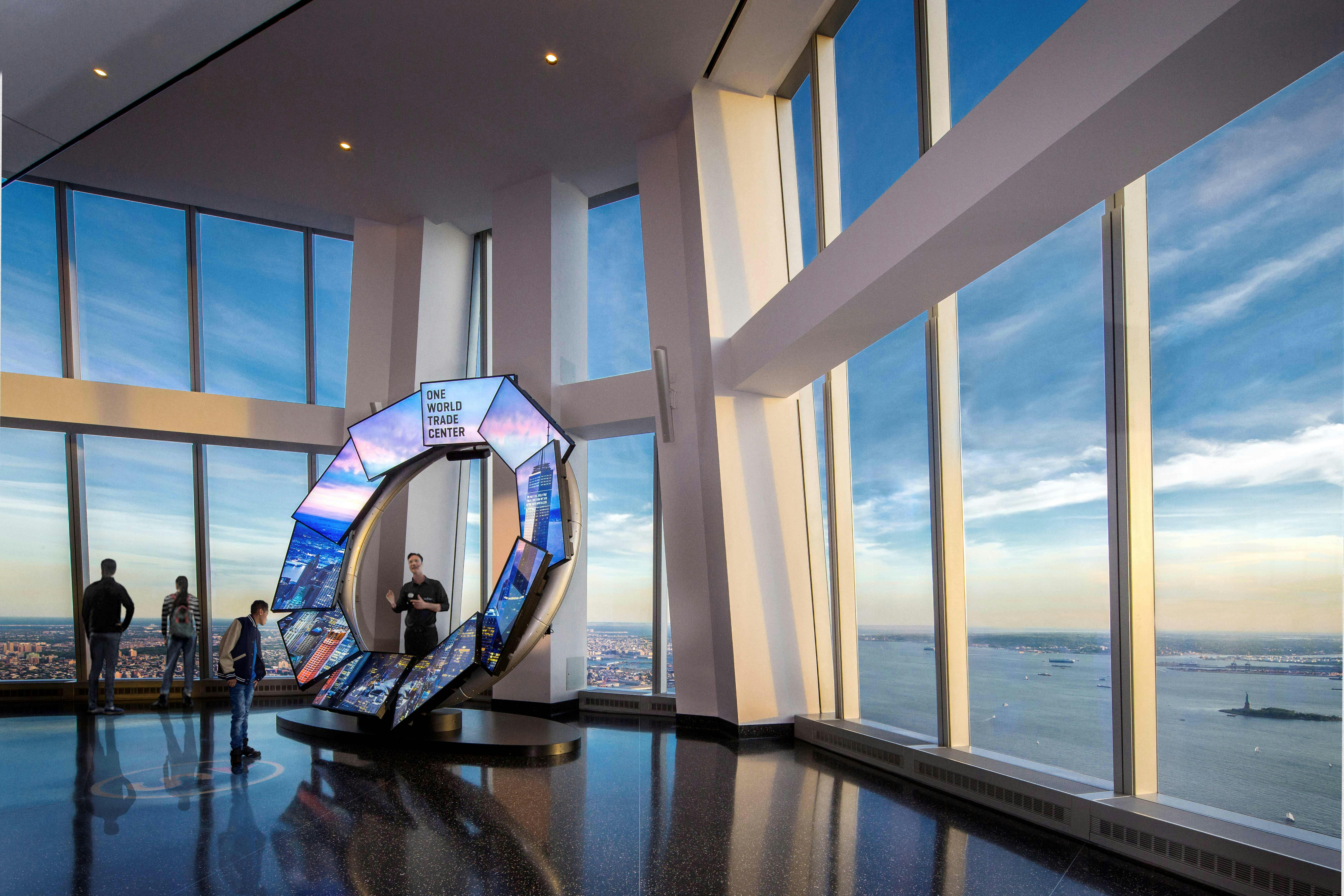 Tall windows showcase a skyline and ocean view. An exhibit with screens and "One World Trade Center" sign stands indoors with two people nearby.