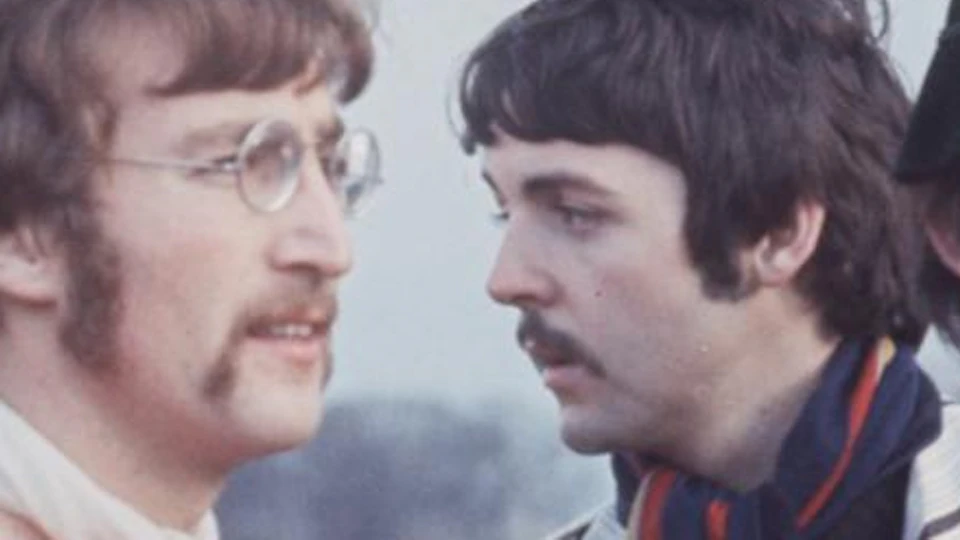 Three men in colorful clothing stand outdoors. Two men in focus, one in a striped jacket and another in a red shirt with a scarf.