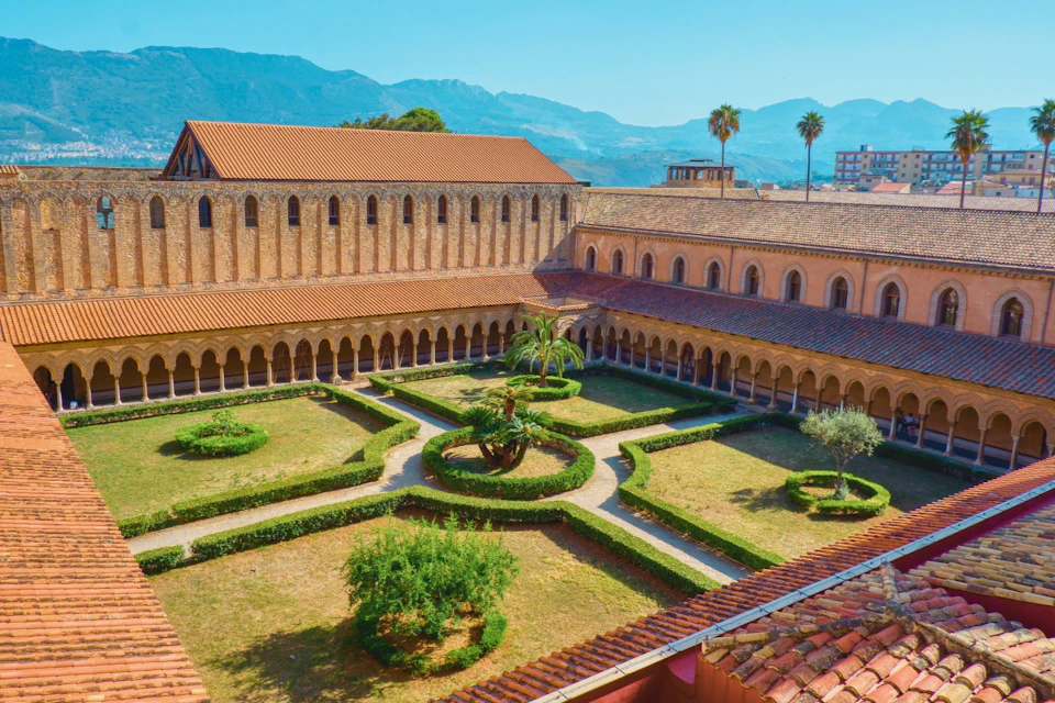 A courtyard with manicured hedges and a central palm tree, surrounded by an arched cloister with mountains in the background.
