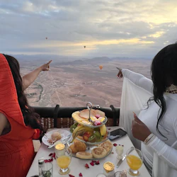 Two people enjoying a meal in a hot air balloon, pointing at other hot air balloons in the distance over a desert landscape.