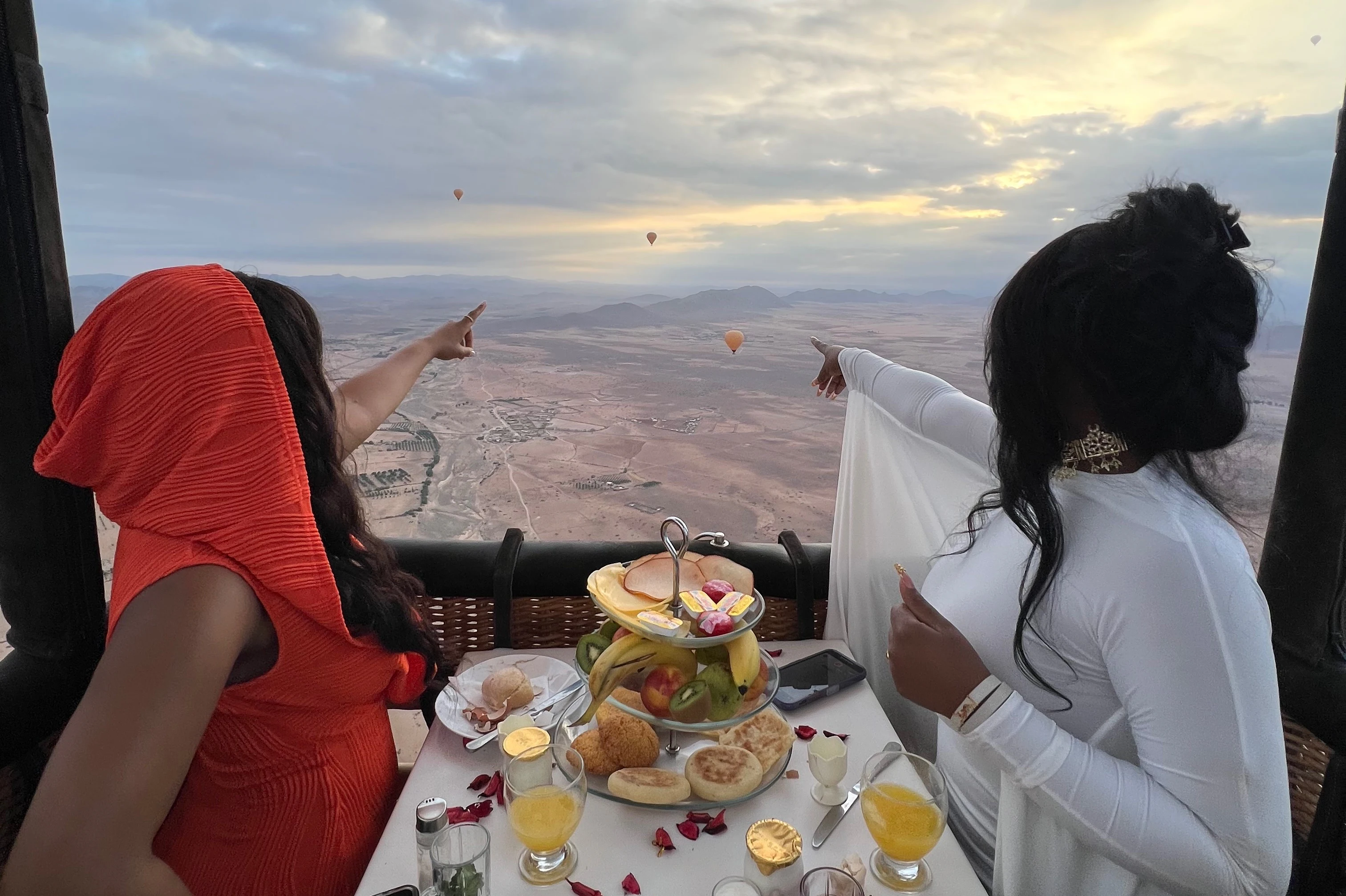 Two people enjoying a meal in a hot air balloon, pointing at other hot air balloons in the distance over a desert landscape.
