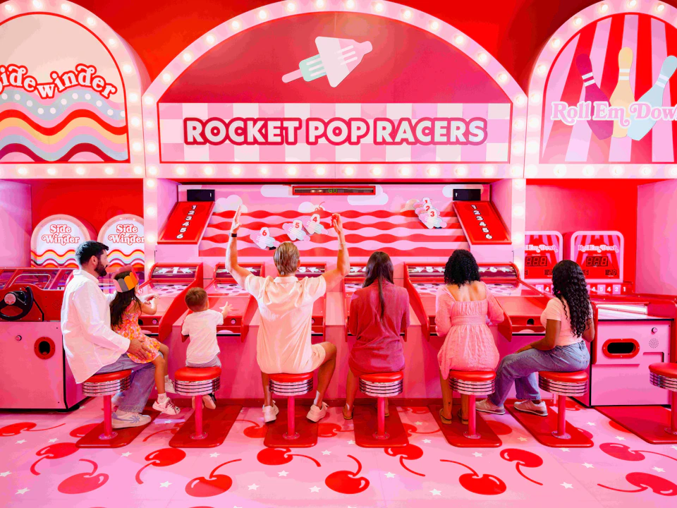 Six people, including two children, on red stools at a Rocket Pop Racers arcade game in a brightly colored, cherry-themed room.