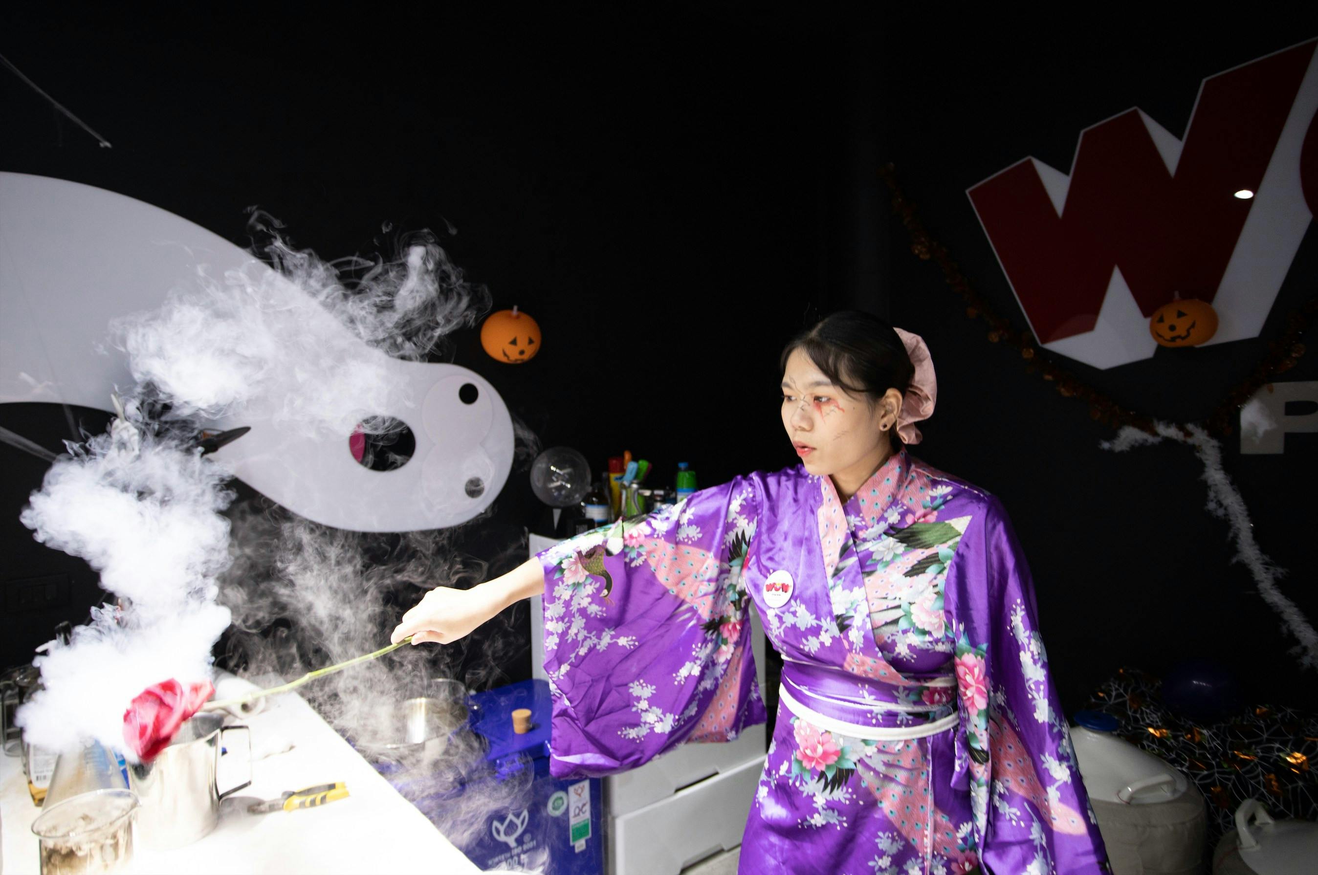 A person wearing a purple kimono in a decorated room with Halloween elements and a dark background.