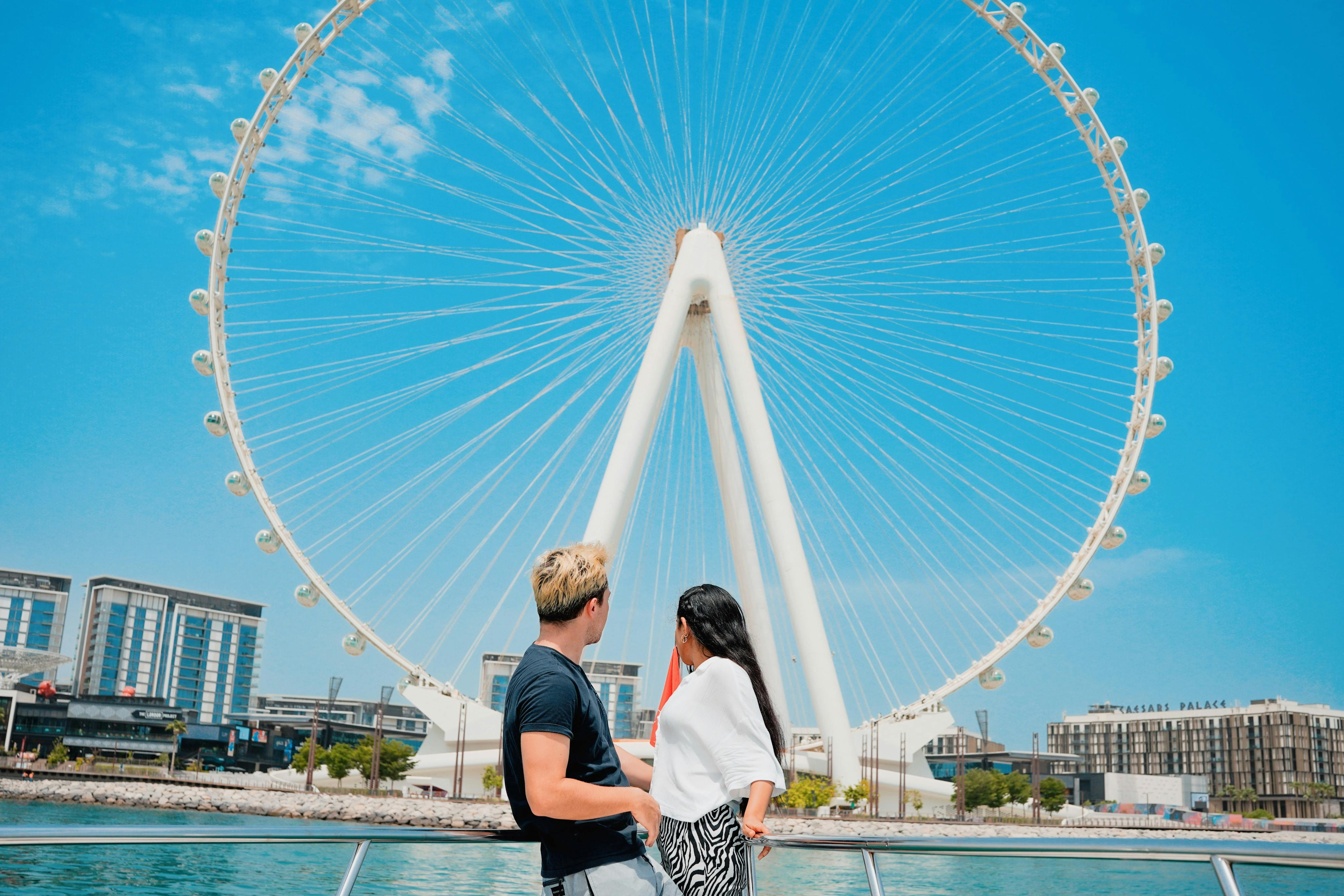 Muž a žena stojí na lodi a dívají se na velké ruské kolo proti jasně modré obloze.