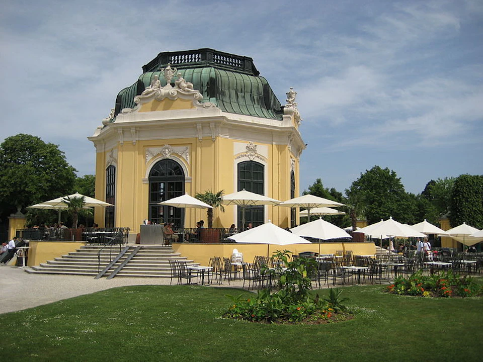 Yellow pavilion with a green dome roof, surrounded by outdoor seating and white umbrellas, set in a green landscaped area.