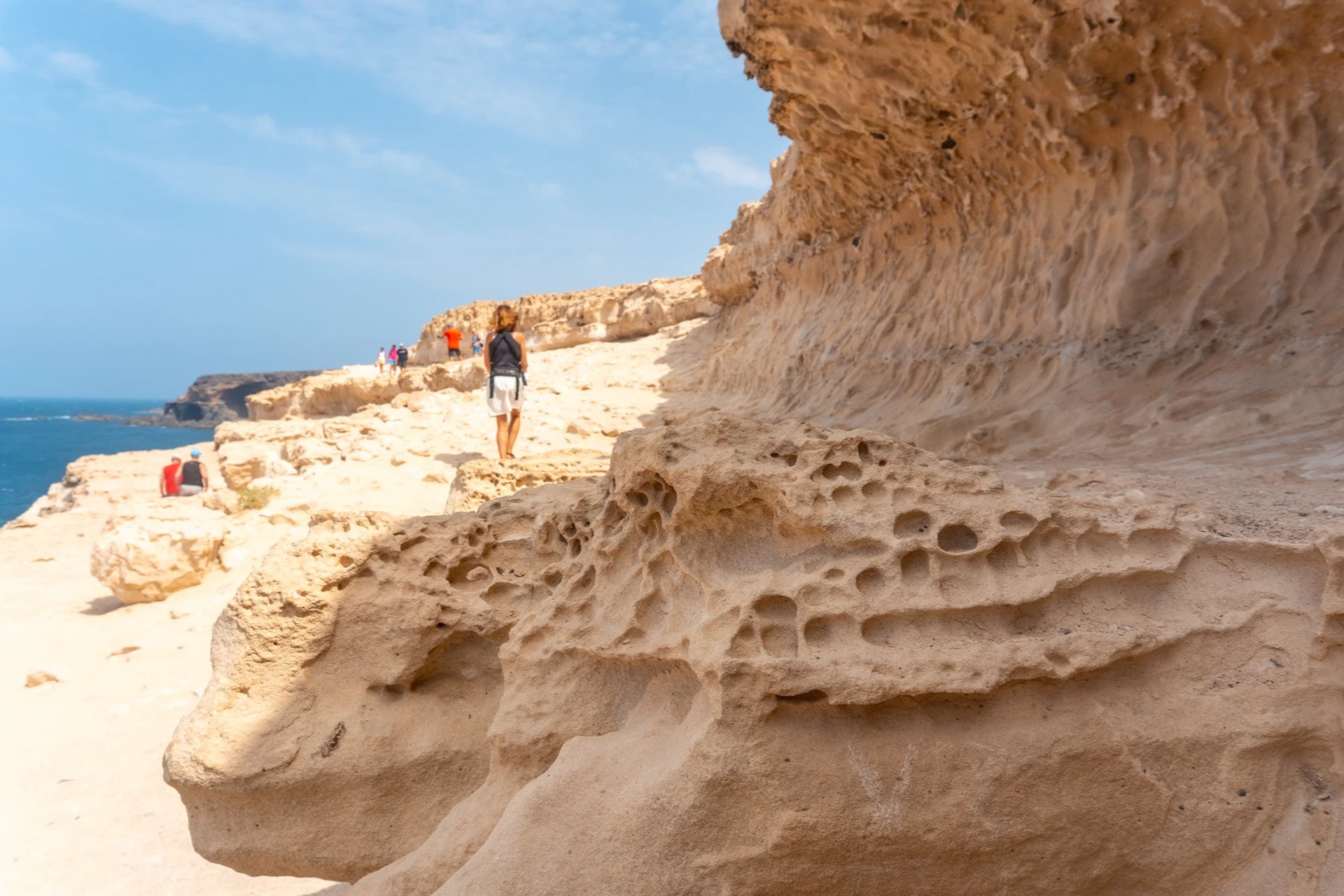 A person wearing a backpack walks along a rocky, eroded cliff path under a clear blue sky, with others in the distance.