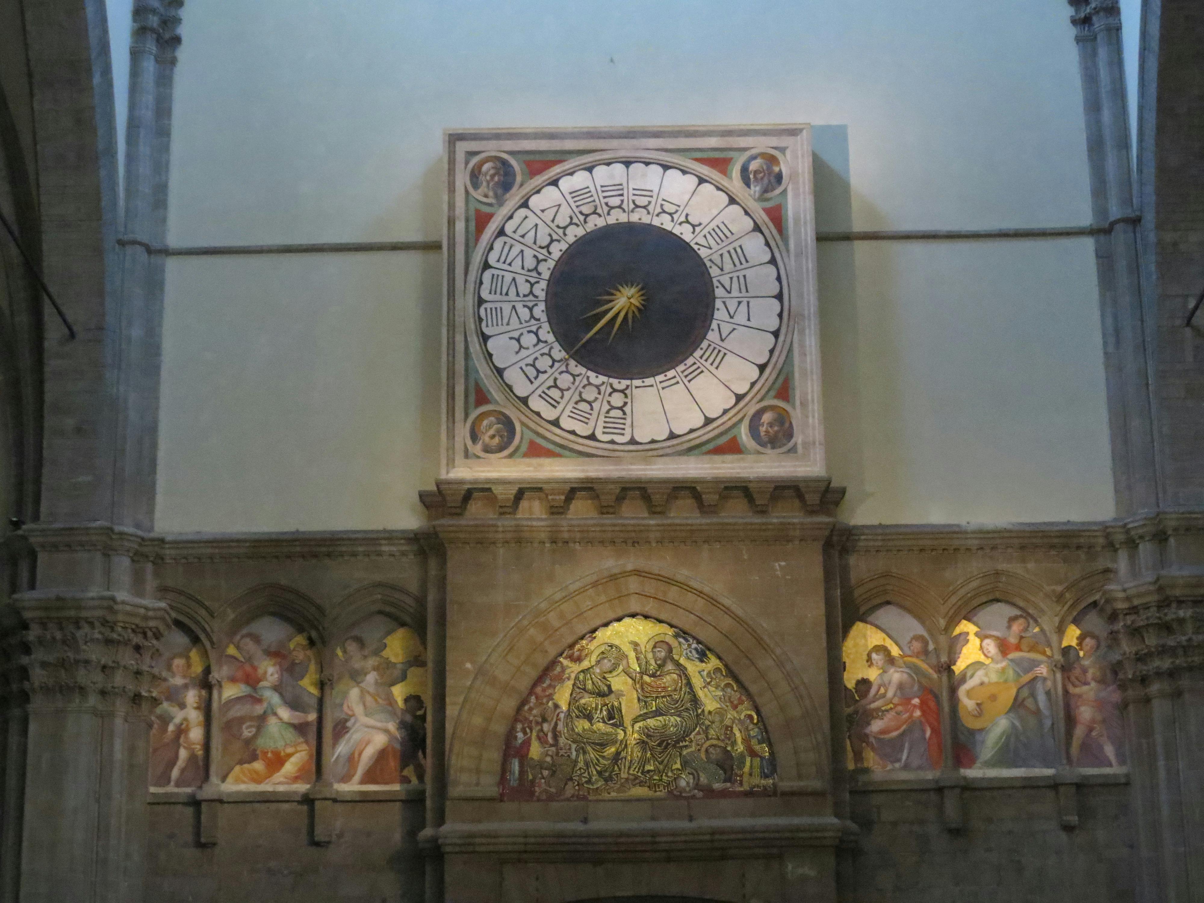 在拱形壁龛上方的教堂内部墙壁上安装了一个带有罗马数字的大型华丽钟表，上面有宗教绘画。