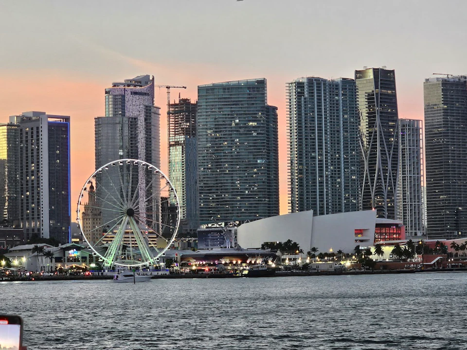 City skyline at dusk featuring a lit Ferris wheel near modern high-rise buildings, with water in the foreground.