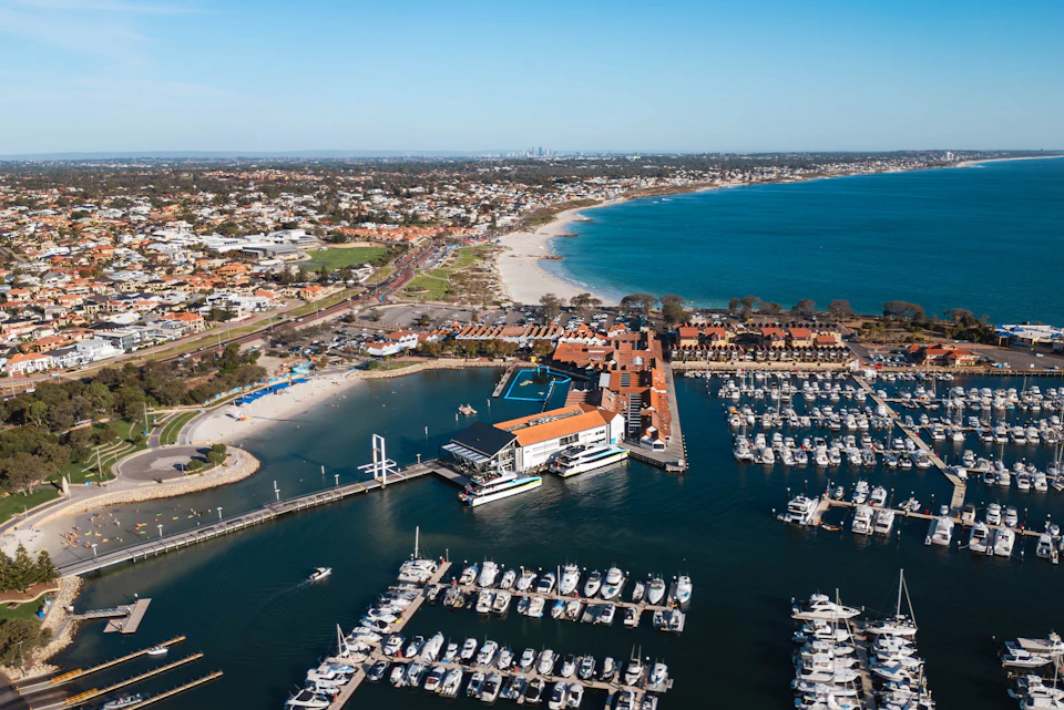 Aerial view of a marina with moored boats, adjacent buildings, and a coastline with a beach and urban area in the background.