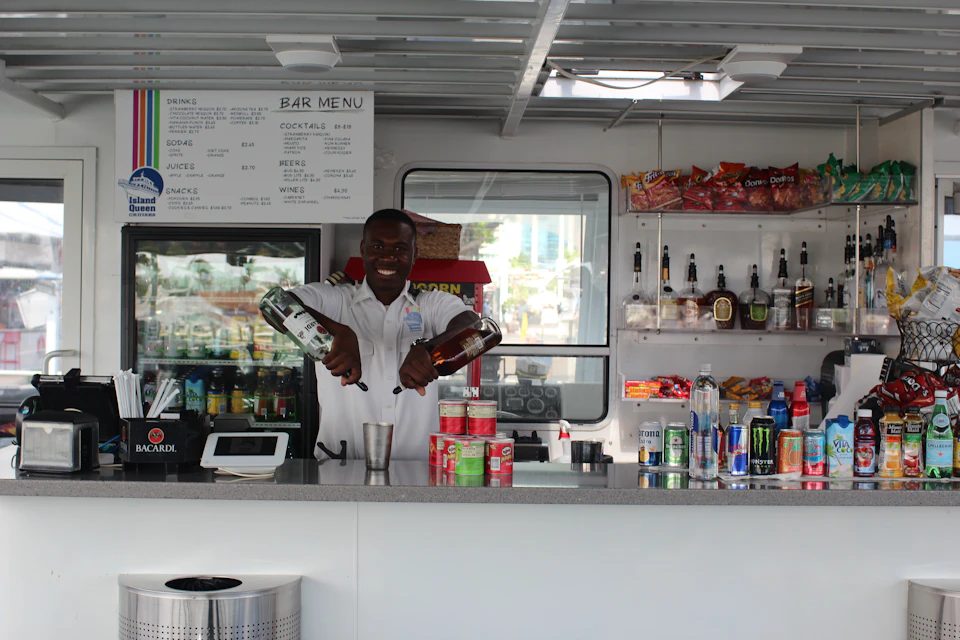 Man in uniform pours drinks at a bar counter with shelves of snacks, bottles, and a menu.
