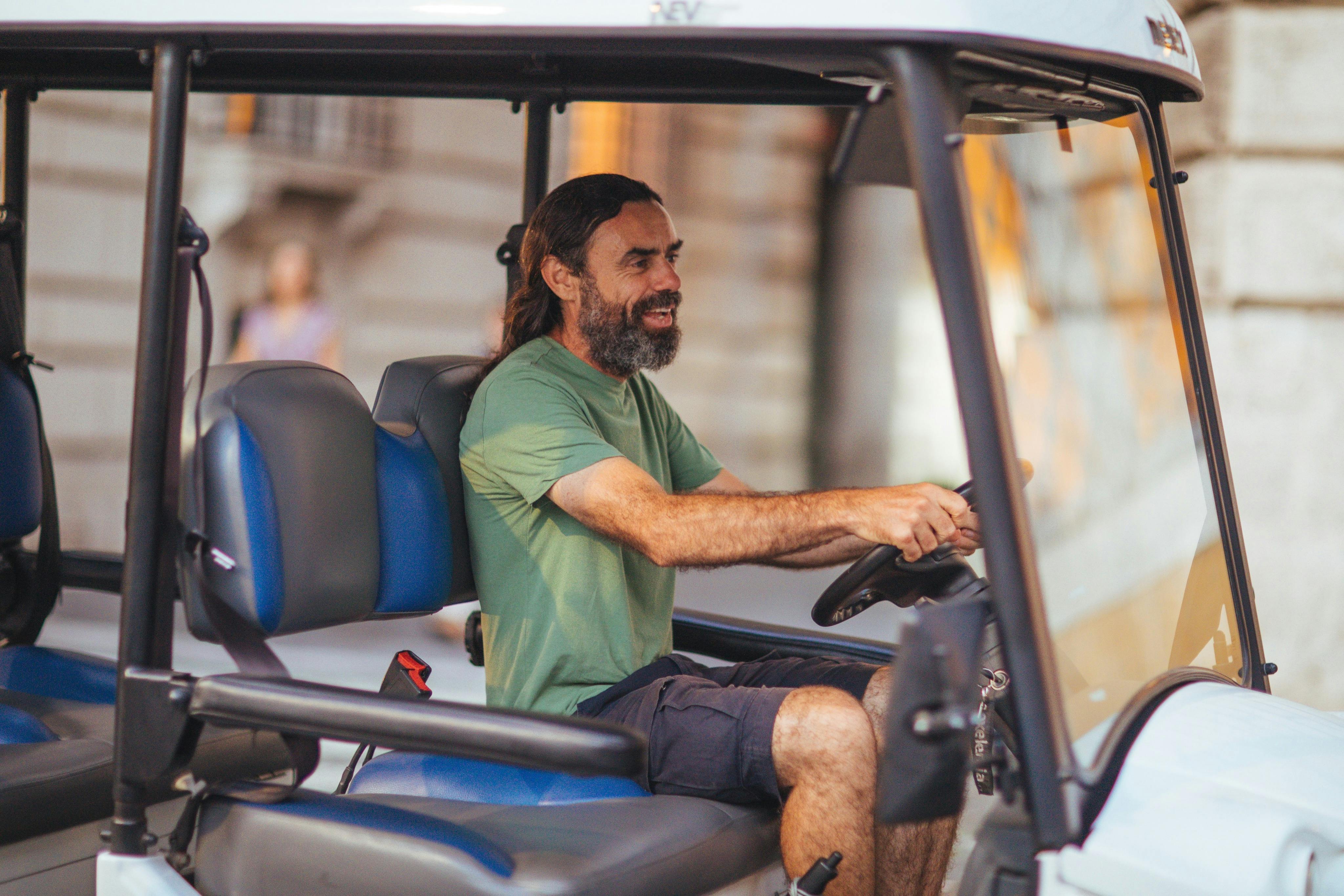 A man with long hair and a beard driving a small open vehicle in an urban setting.