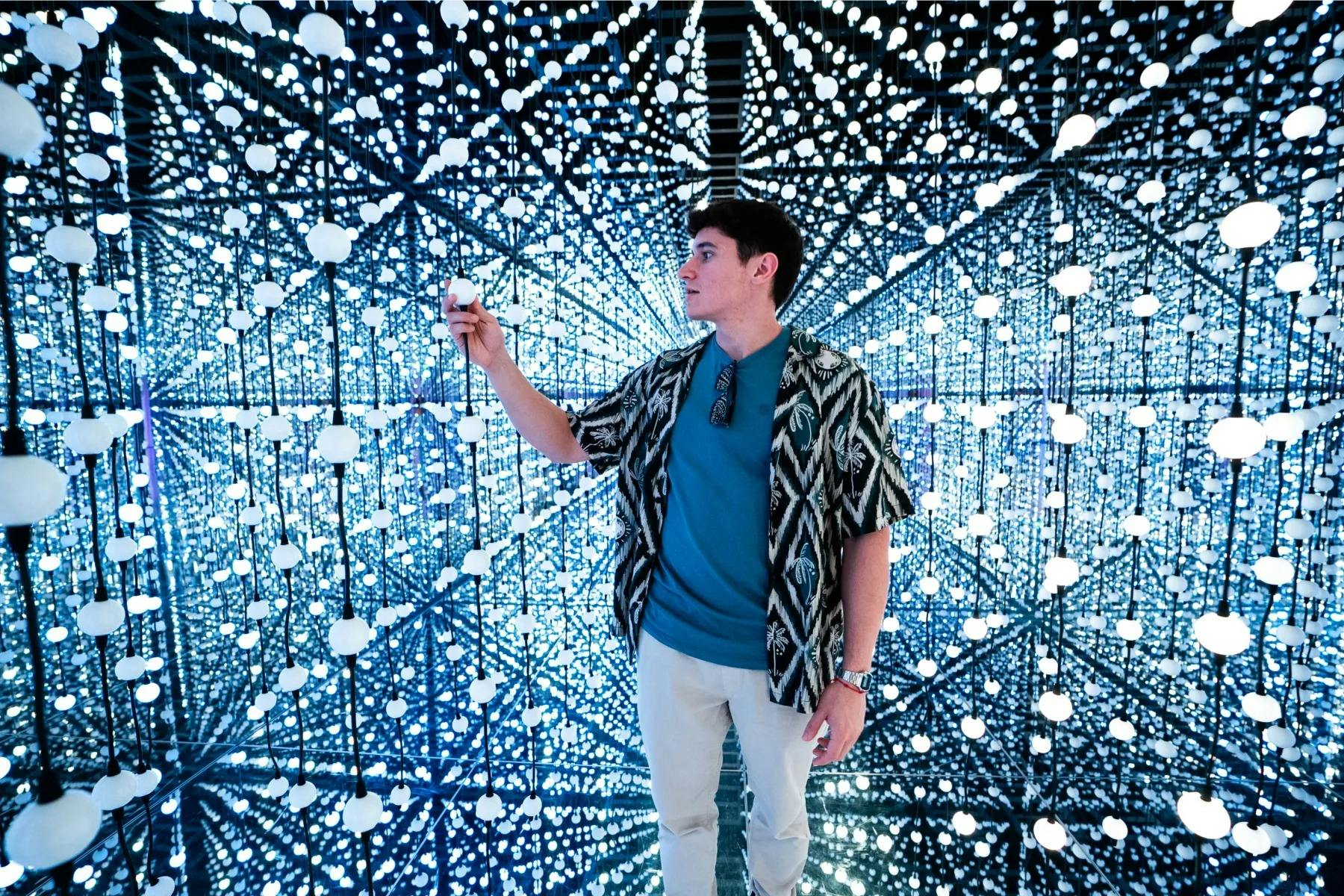 ミラーリングされた幾何学的な部屋の中で、小さな輝く光の中に立っている人物が、その光の1つに触れている。