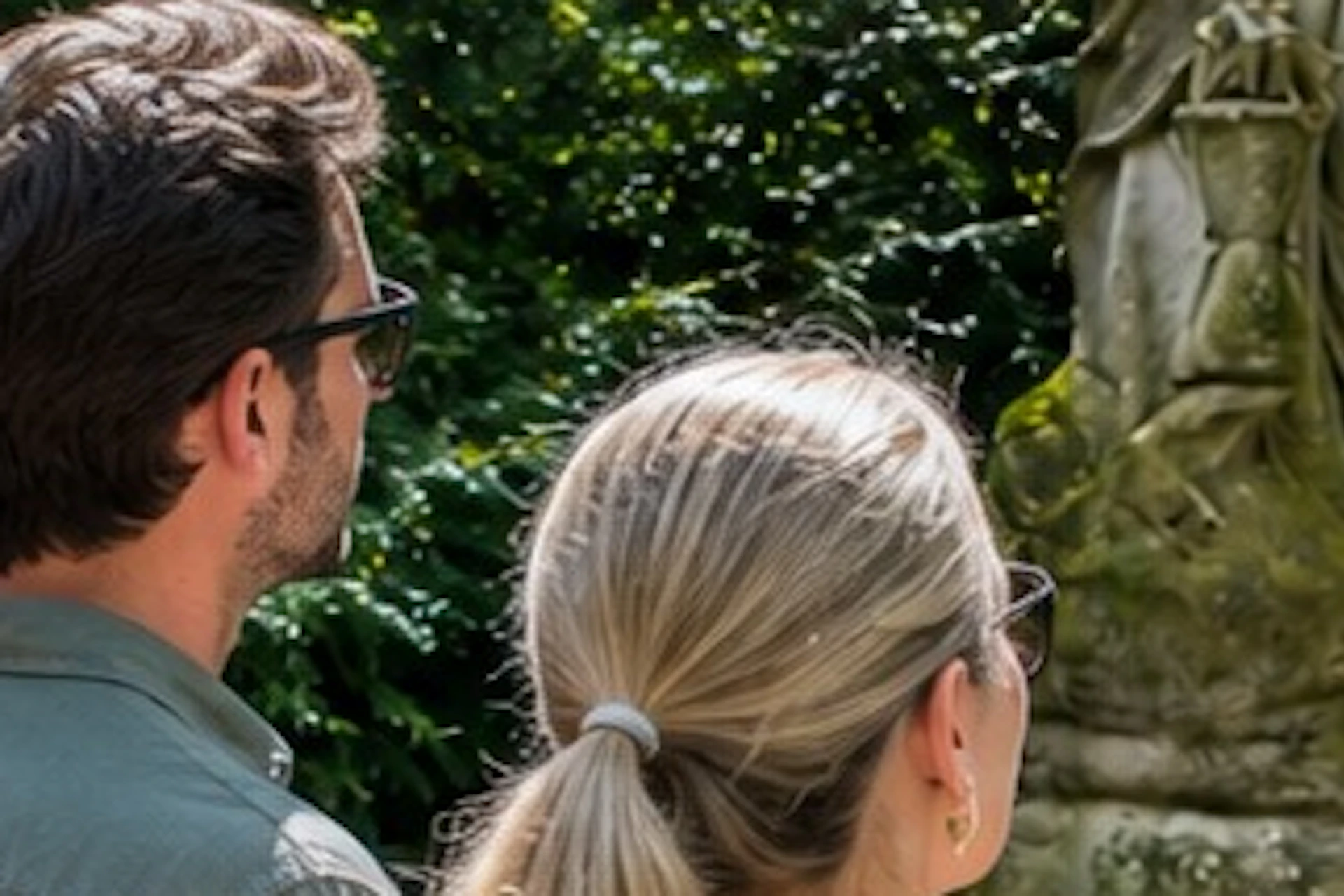 A tour guide is speaking to three visitors in front of a statue of a skeletal figure holding a scythe, set in a lush garden.