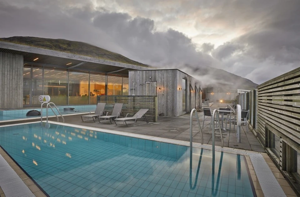 Modern outdoor pool area with reclining chairs, a wooden building, and steam rising. Mountains and cloudy sky in the background.