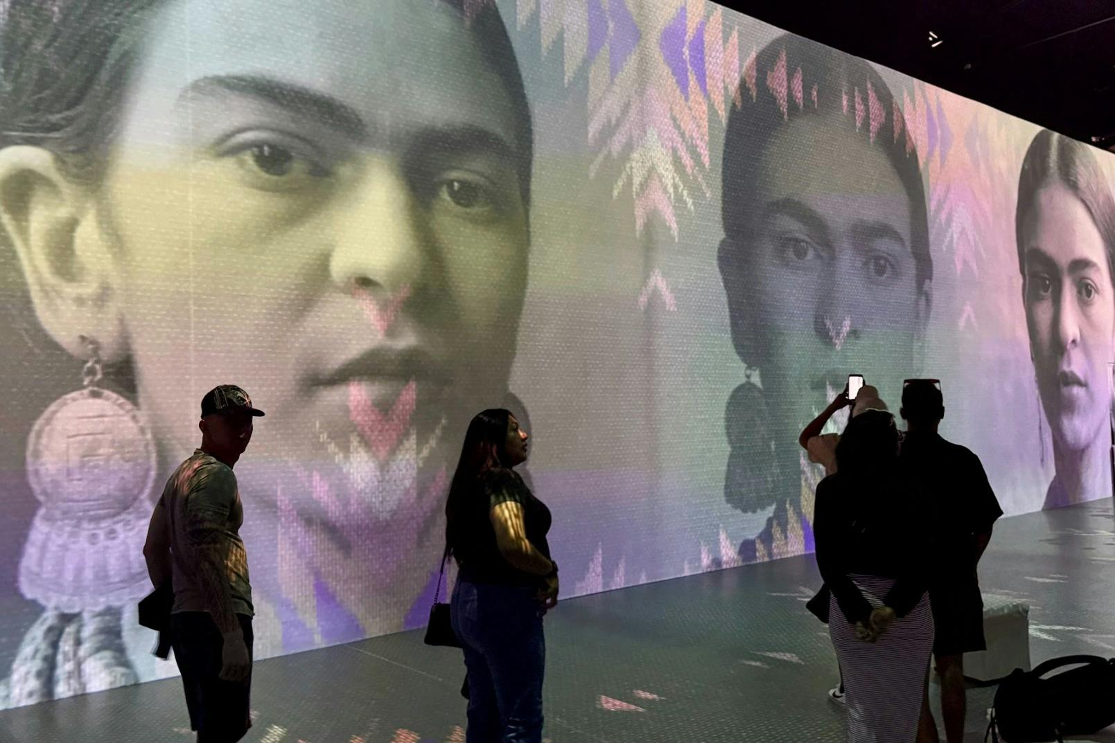 Lidé pozorující velkou nástěnnou projekci s portréty ženy s barevnými symetrickými vzory.