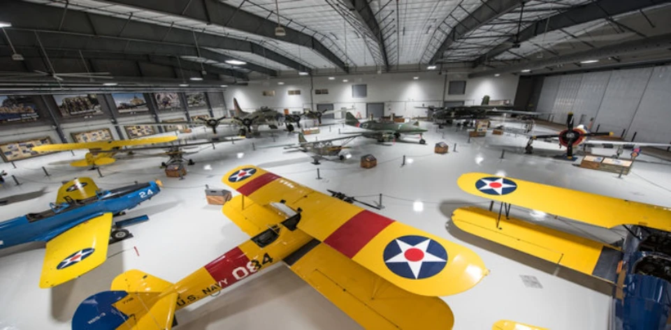 A museum hangar showcasing vintage military aircraft, including a prominent yellow biplane in the foreground with U.S. Navy markings.