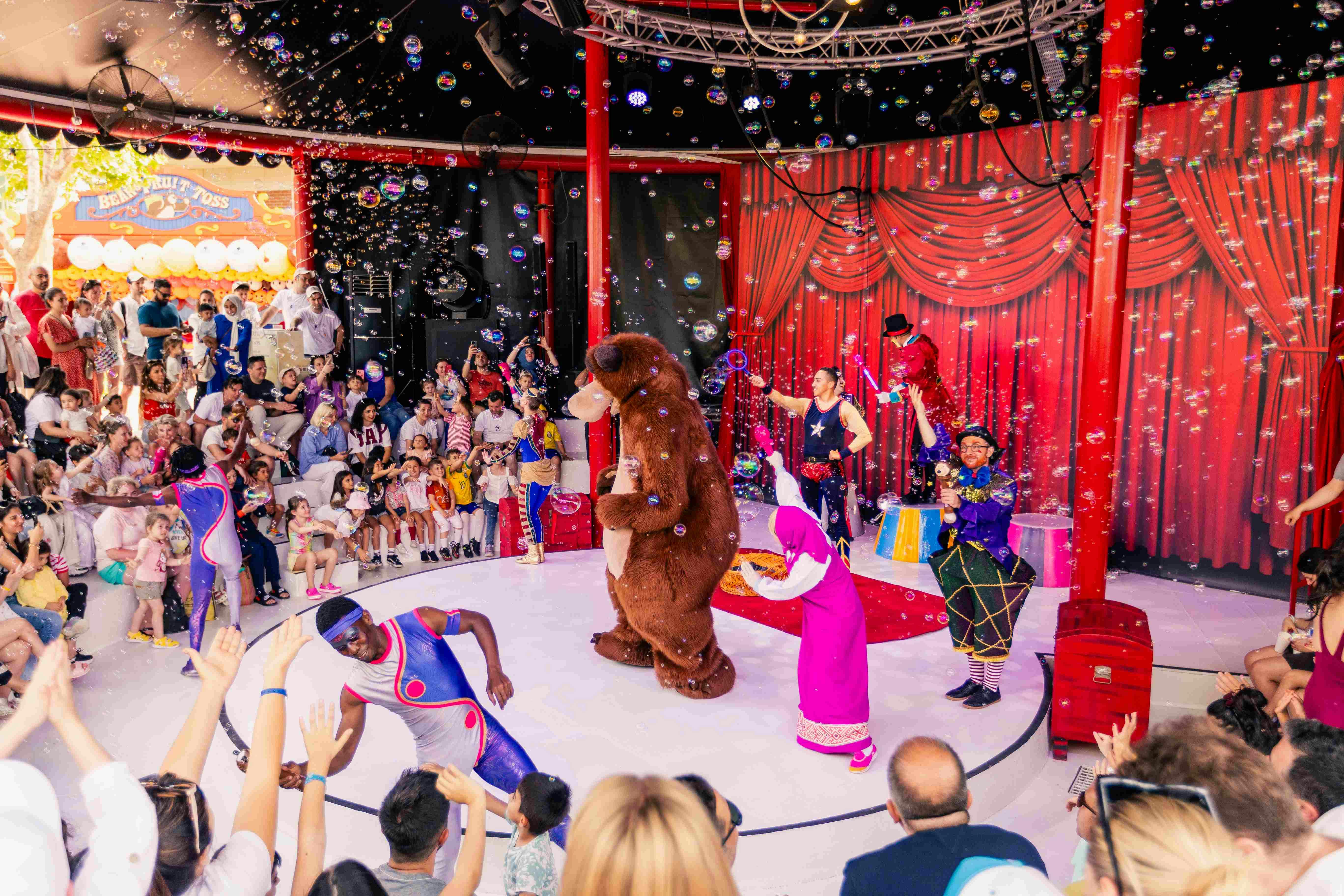 Performers in colorful costumes, including a bear, entertain a crowd in a circus-themed setting with bubbles in the air.