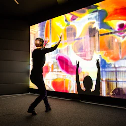 En person interagerar med en färgglad, abstrakt digital display och kastar skuggor med upplyfta händer i ett mörkt rum.