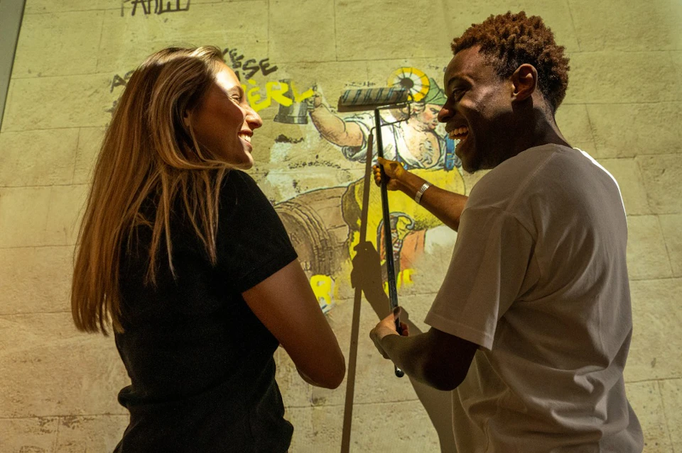 A woman with long blonde hair observes a person using a roller brush to apply artwork to a wall.